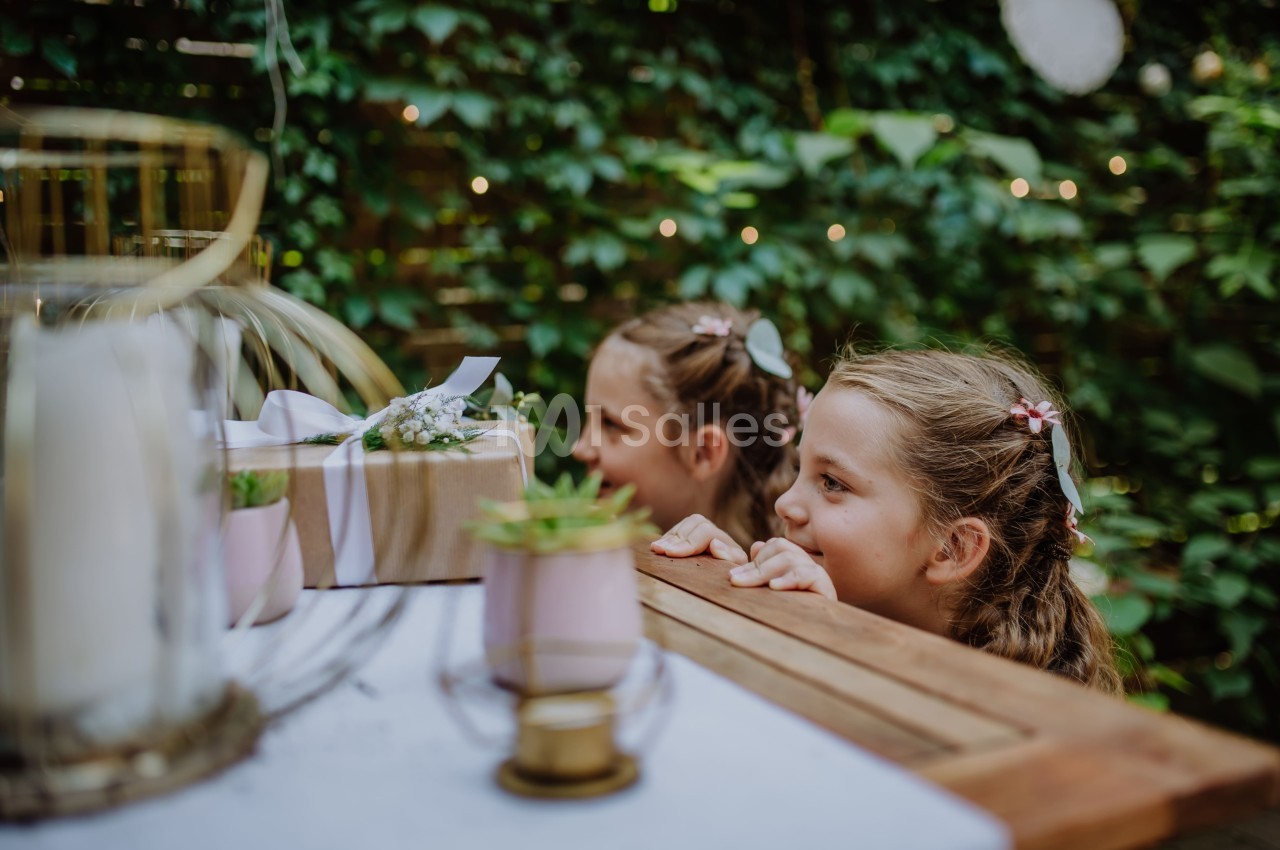 Deux enfants souriants regardent des cadeaux sur une table en bois, dans un jardin décoré de plantes et de lumières.