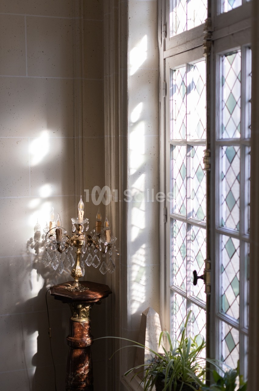 Lumière naturelle traversant une fenêtre à vitraux, éclairant un chandelier en cristal sur un pied en marbre.