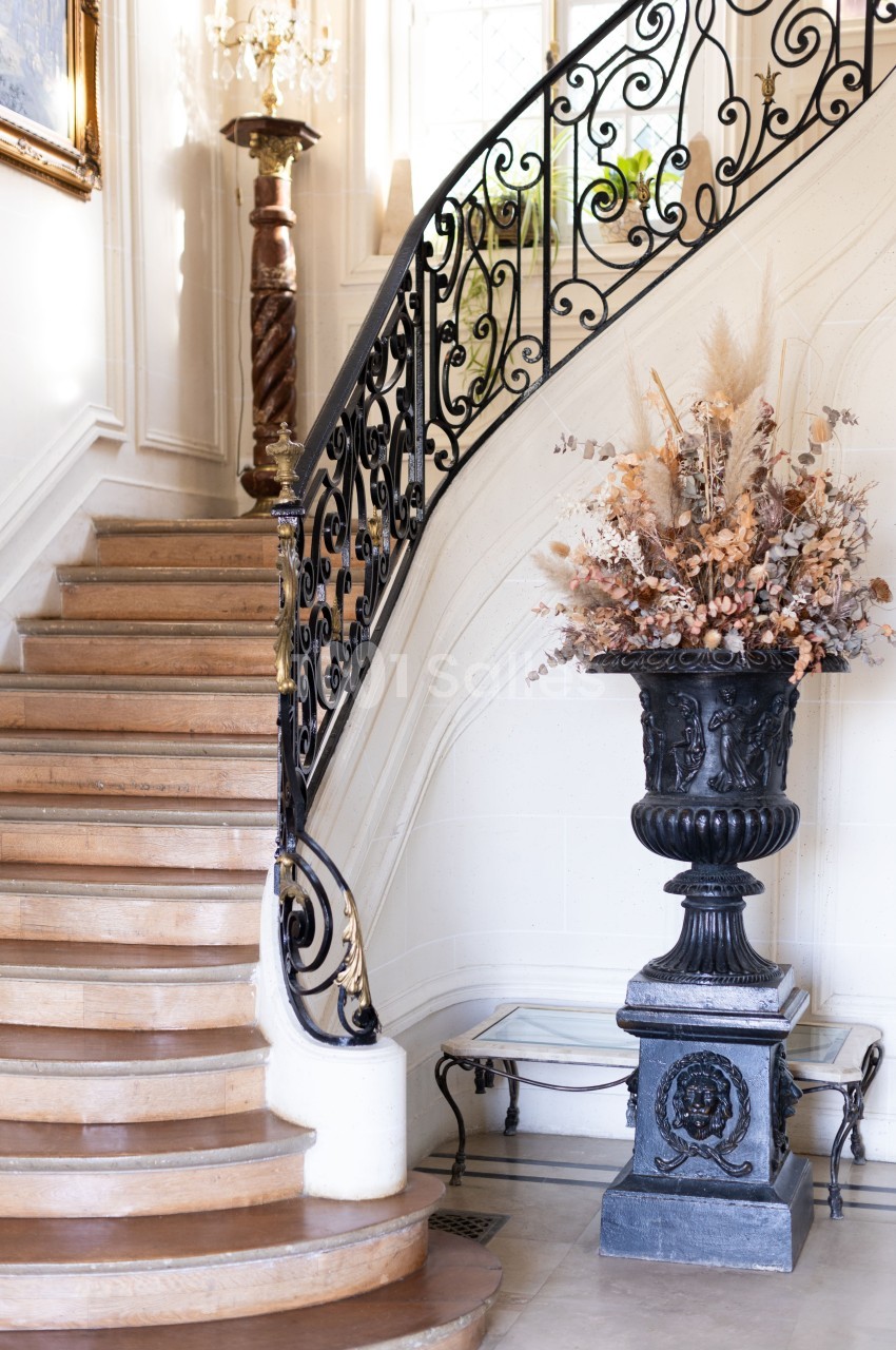 Escalier en bois avec rampe en fer forgé, grand vase décoratif et lumière naturelle entrant par une fenêtre.