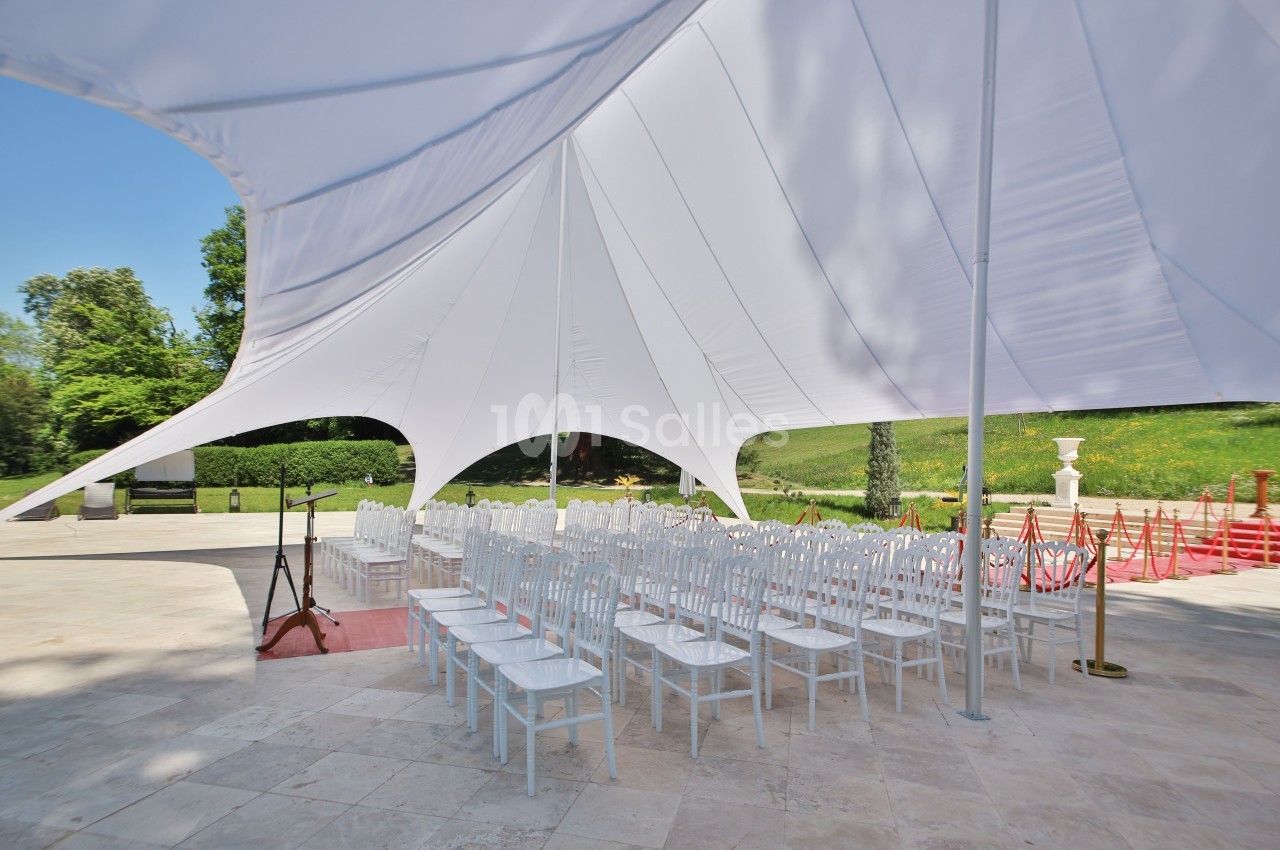 Chaises blanches alignées sous une grande tente blanche dans un espace extérieur pavé, entouré de verdure.
