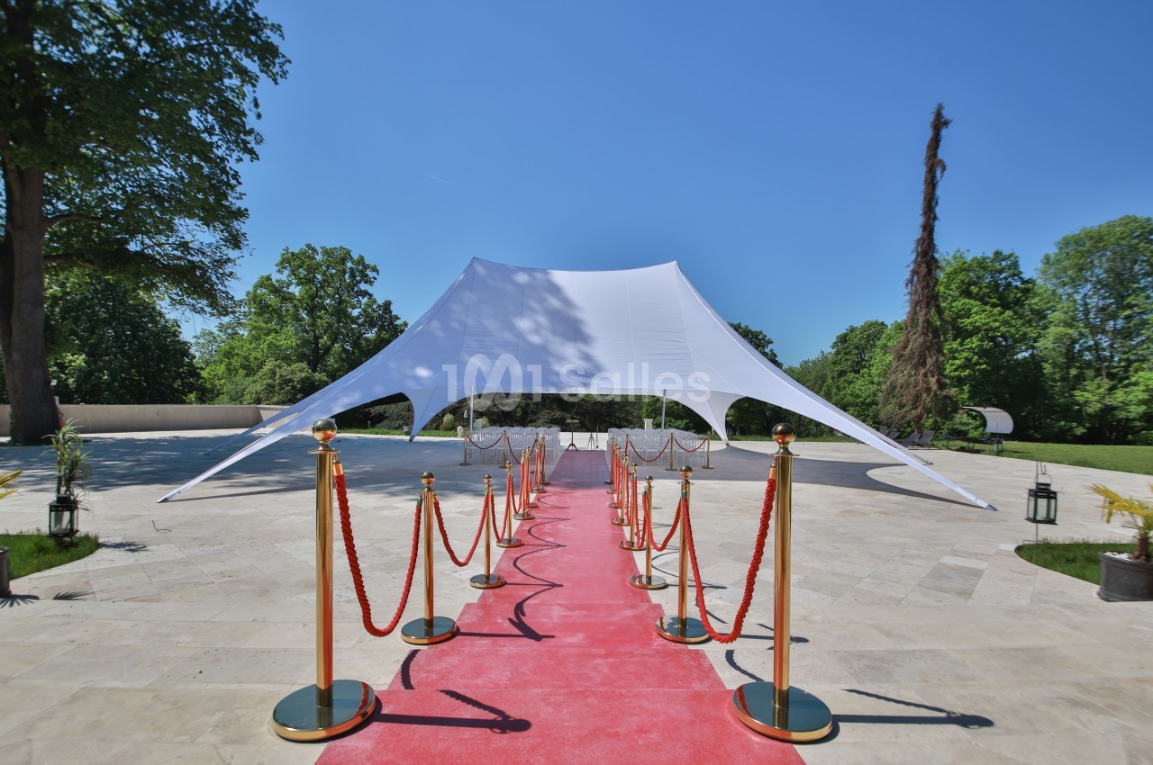 Tente blanche tendue sur une esplanade pavée, avec tapis rouge bordé de poteaux dorés et cordons rouges.
