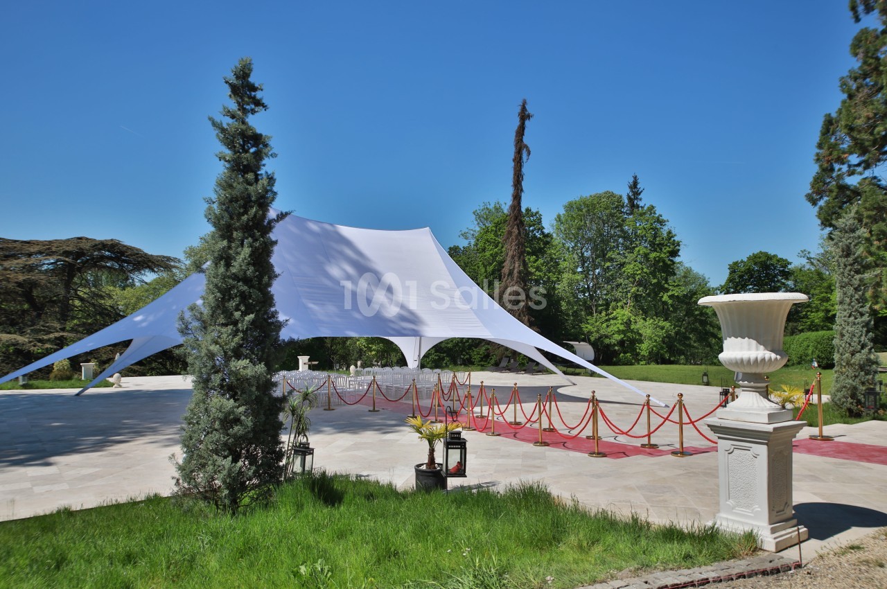 Tente blanche tendue sur une esplanade pavée, entourée de verdure et délimitée par des cordons rouges.