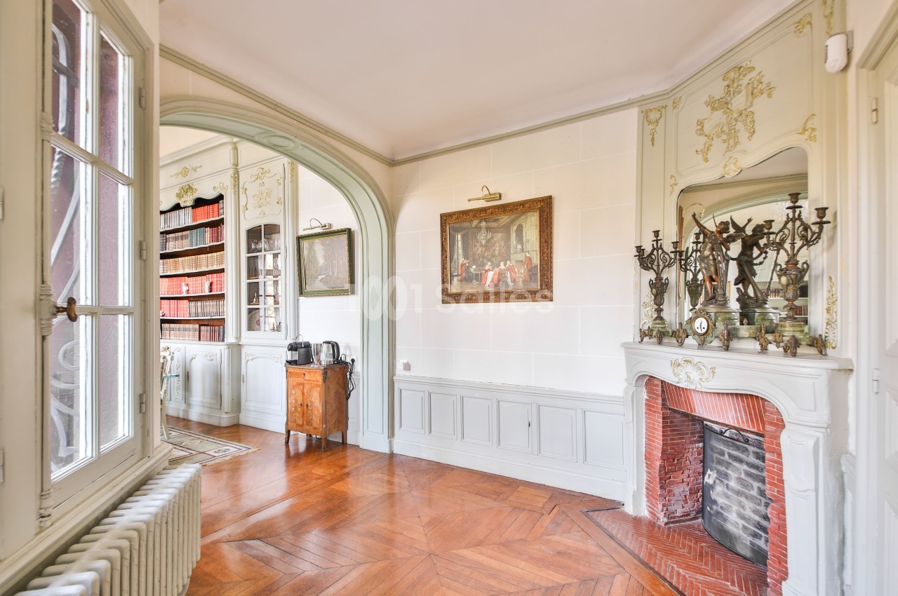 Intérieur lumineux avec parquet, cheminée ornée, bibliothèque et moulures décoratives sur les murs.