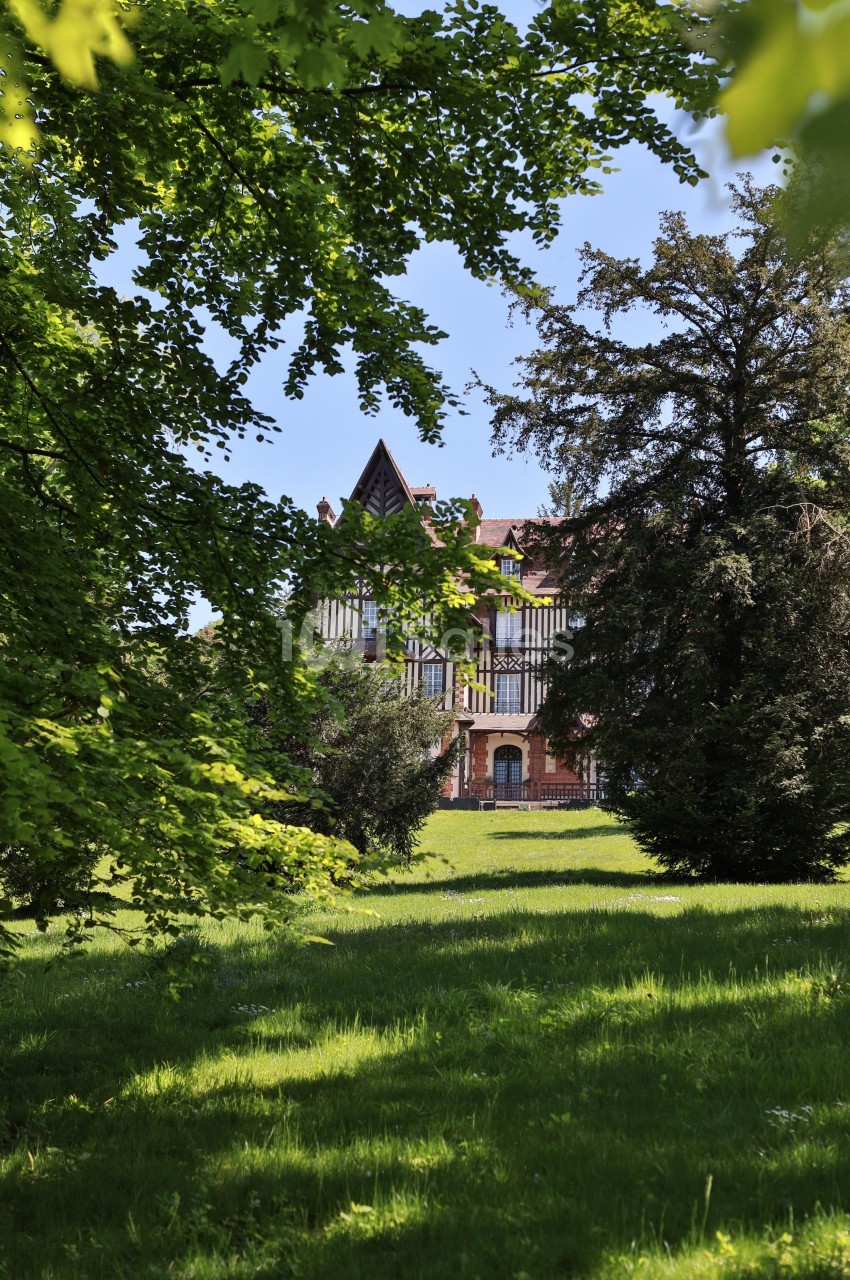 Manoir à colombages entouré d'arbres et d'une pelouse verdoyante sous un ciel bleu.