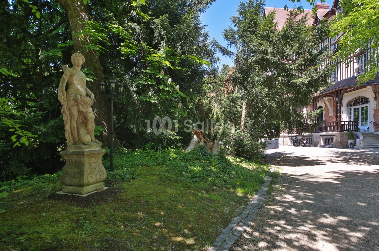 Statue en pierre dans un jardin boisé, près d'une allée menant à une grande maison à colombages.