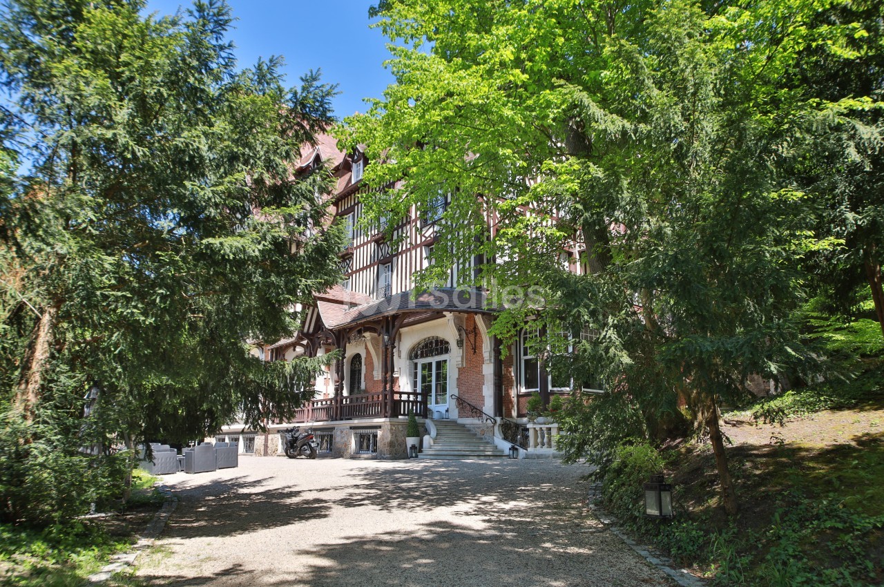 Façade d'une grande maison à colombages entourée d'arbres, avec une allée gravillonnée menant à l'entrée.