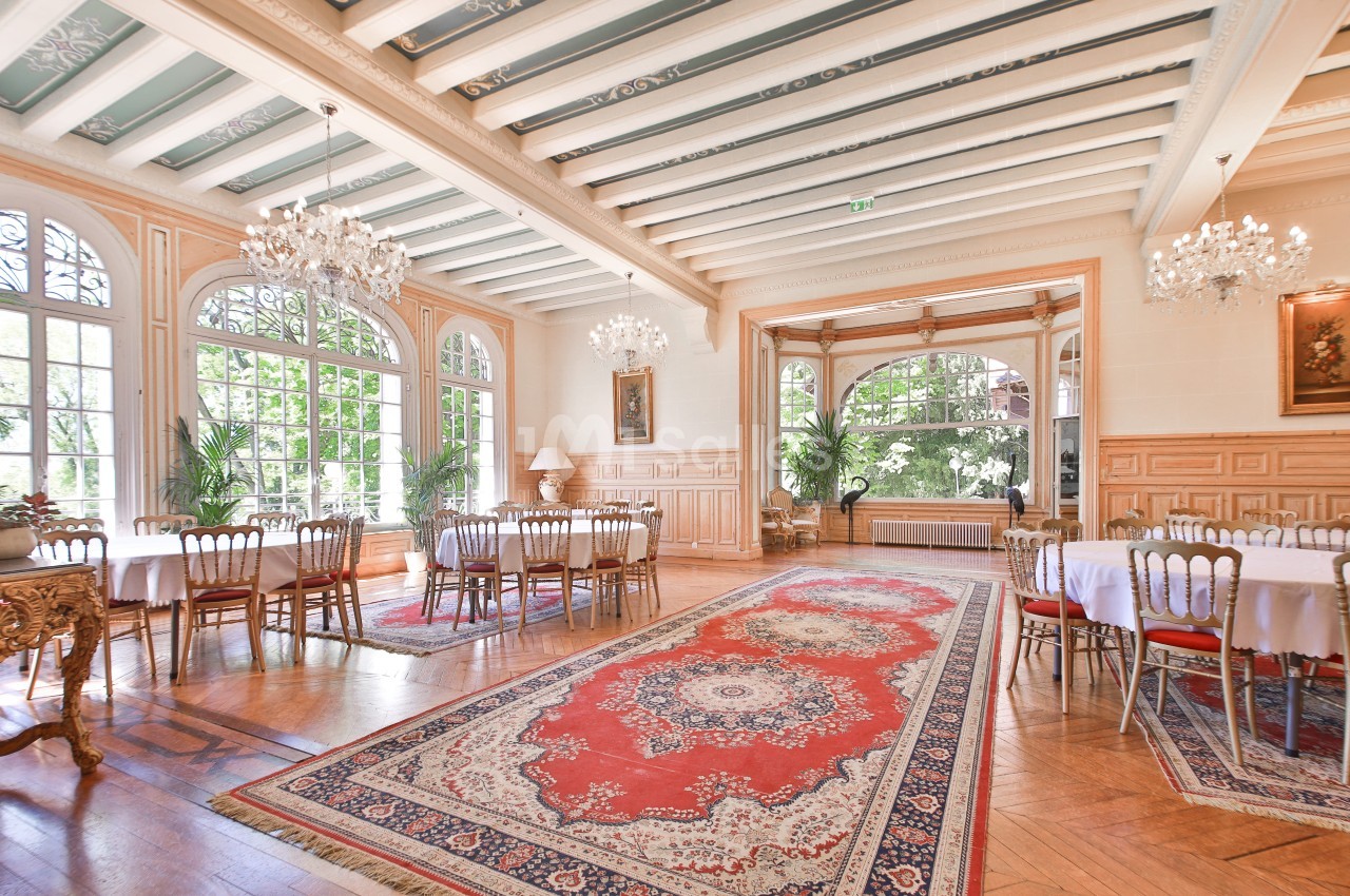 Salle lumineuse avec grandes fenêtres, lustres, tables rondes dressées, tapis rouge et parquet en bois clair.