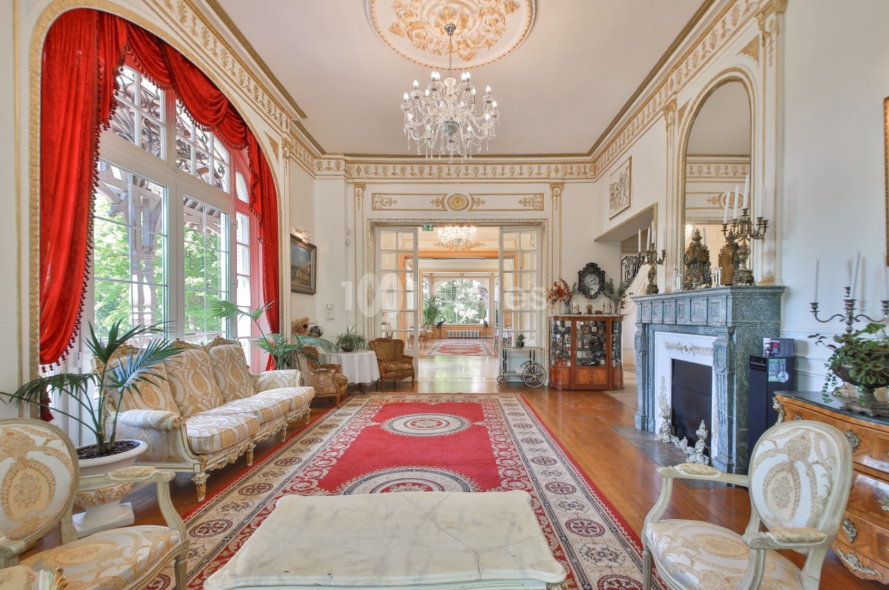 Salon élégant avec mobilier classique, cheminée en marbre, lustres, tapis rouges et grandes fenêtres donnant sur un jardin.