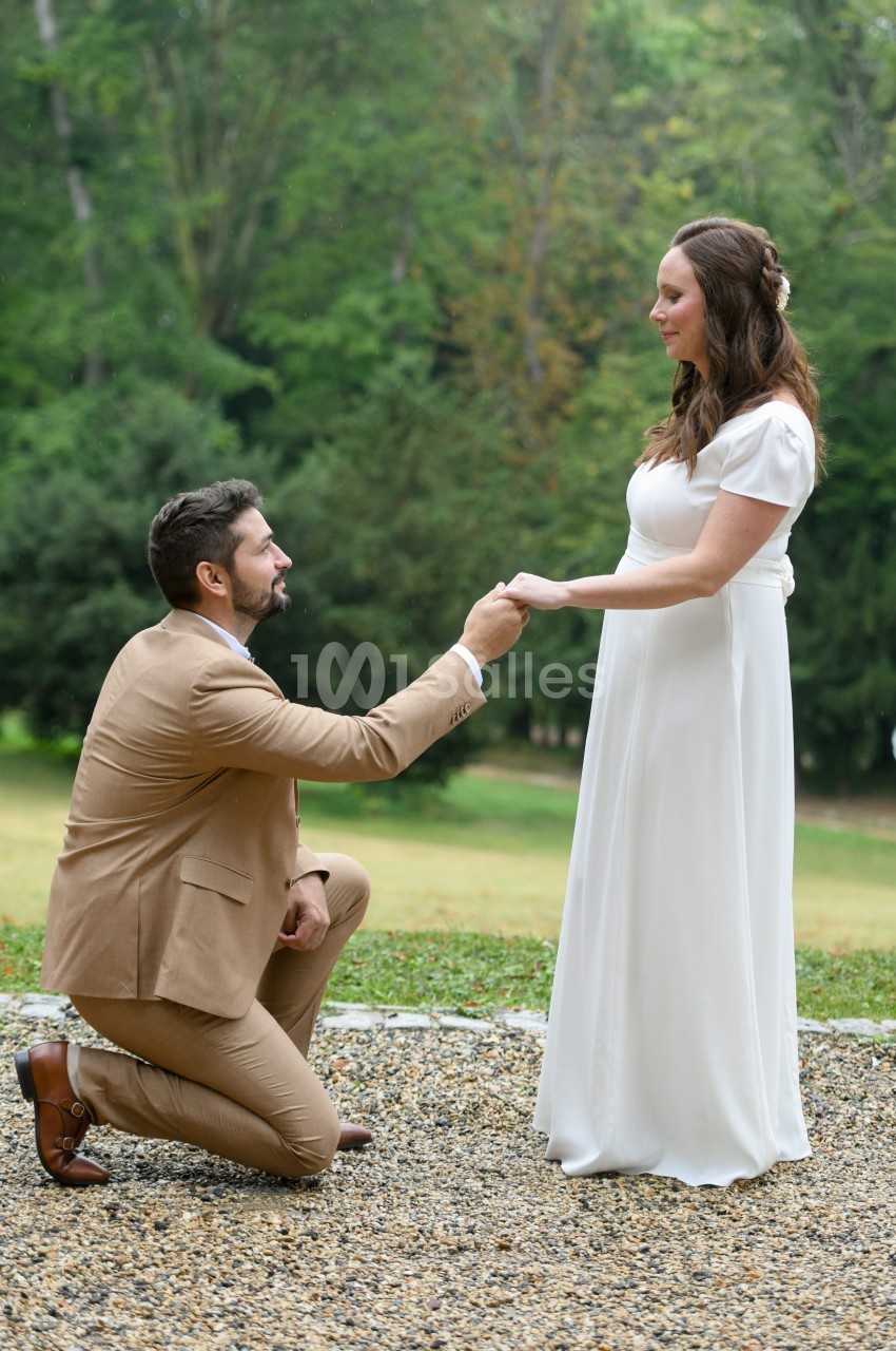 Un homme en costume beige, à genoux, tient la main d'une femme en robe blanche dans un parc verdoyant.