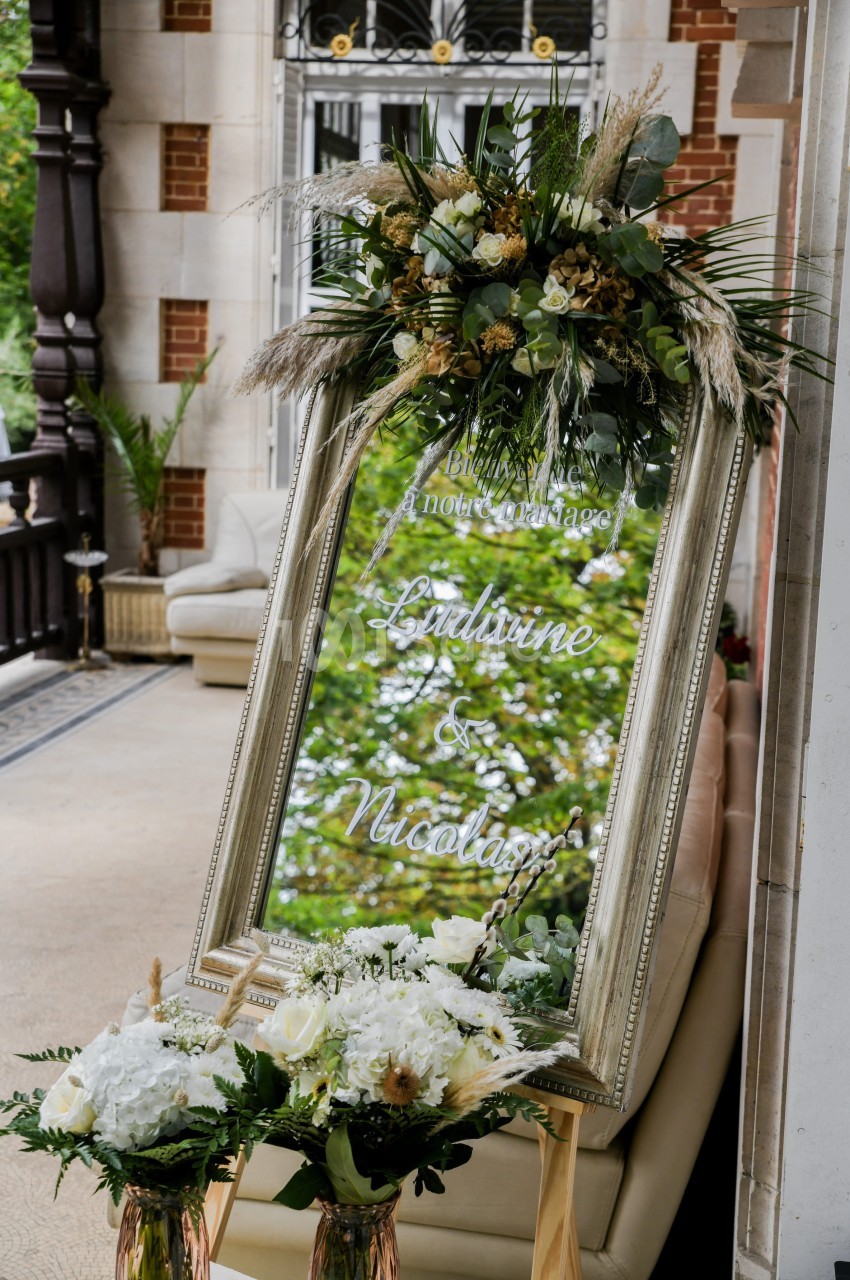 Miroir décoratif avec fleurs et feuillages, affichant des noms gravés, placé sur une terrasse élégante.