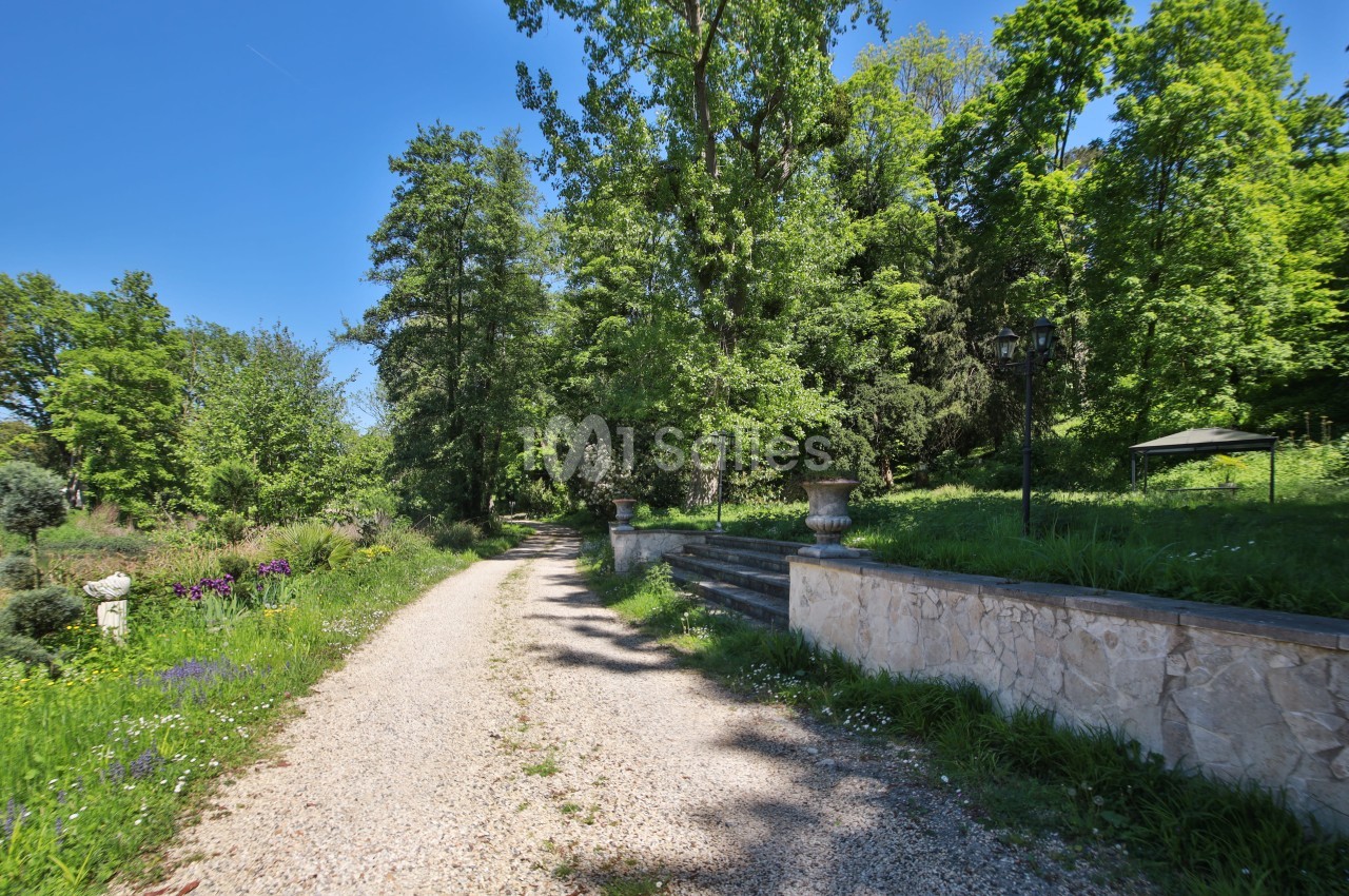 Chemin de gravier bordé de verdure et d'arbres, avec un muret en pierre et des marches sur la droite.