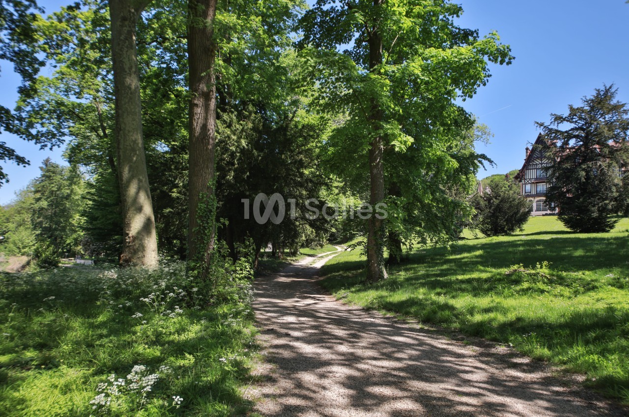 Chemin ombragé traversant un parc verdoyant avec des arbres et une maison visible en arrière-plan.