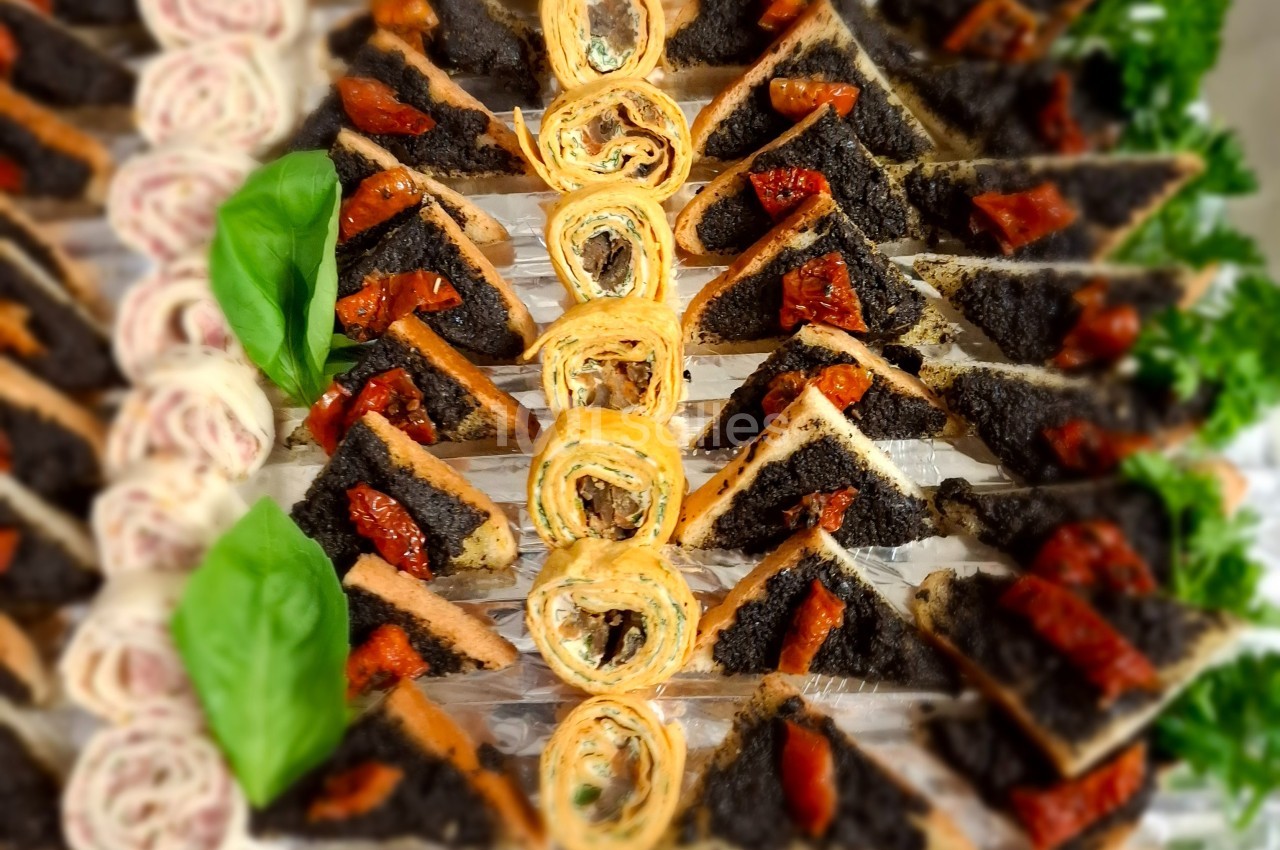 Plateau de toasts triangulaires à la tapenade et tomates séchées, entourés de roulés apéritifs et feuilles de basilic.