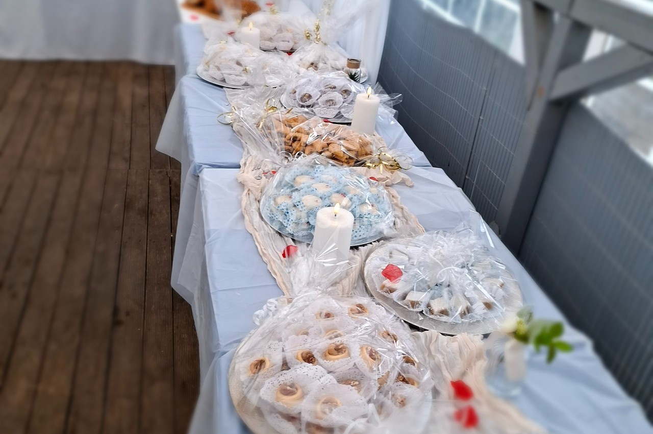 Table garnie de pâtisseries emballées, décorée de bougies blanches et de nappes claires sur une terrasse en bois.