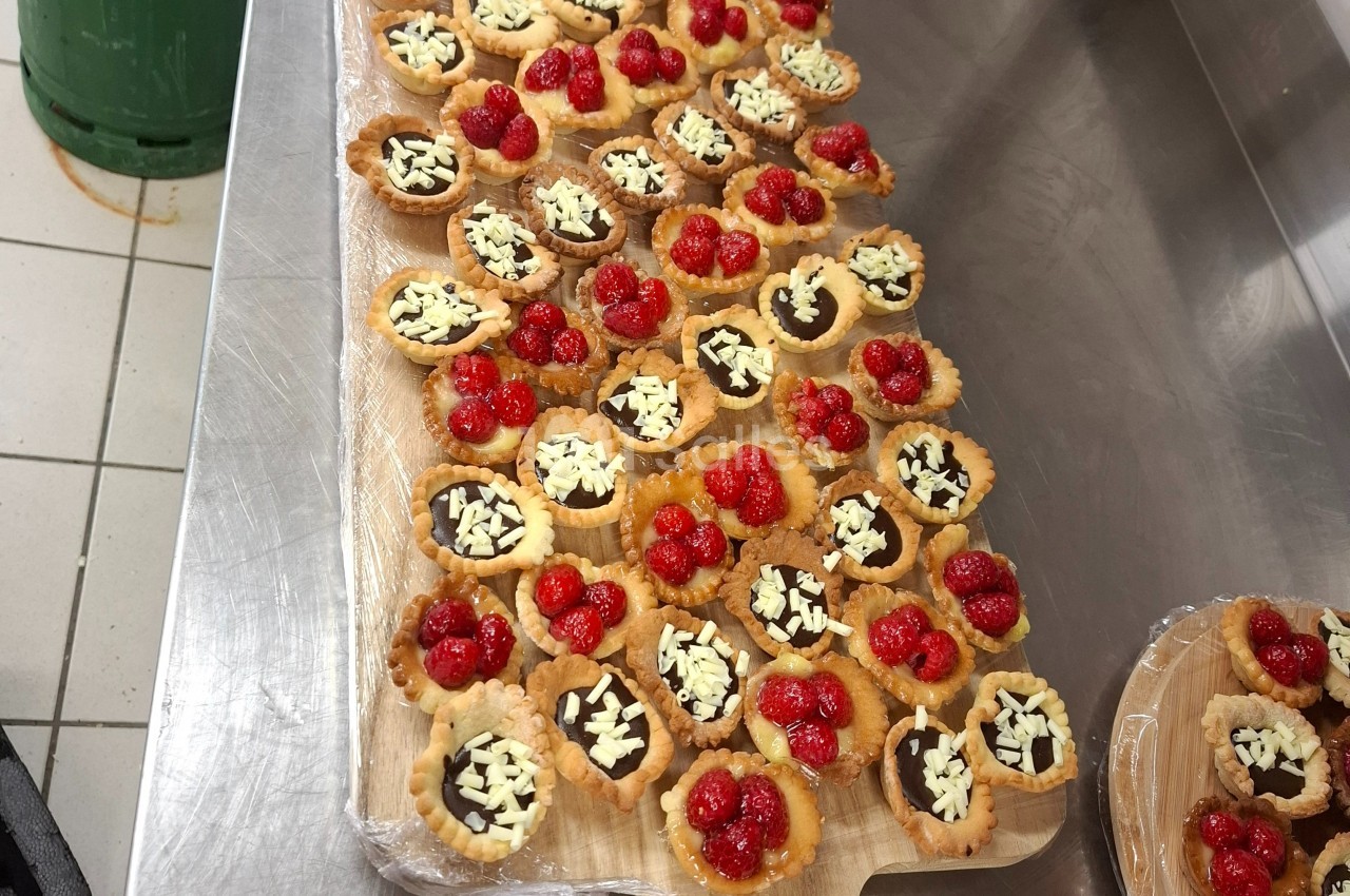 Plateau de tartelettes garnies de chocolat, éclats de chocolat blanc et framboises, posé sur un plan de travail en inox.