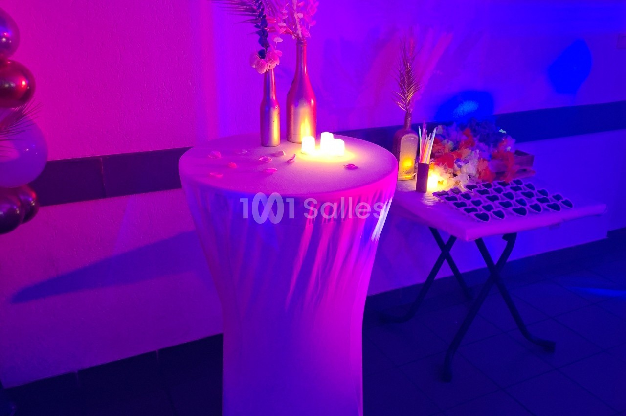 Table haute décorée avec des bougies, fleurs et bouteilles, éclairée par une lumière colorée, avec une table annexe garnie.