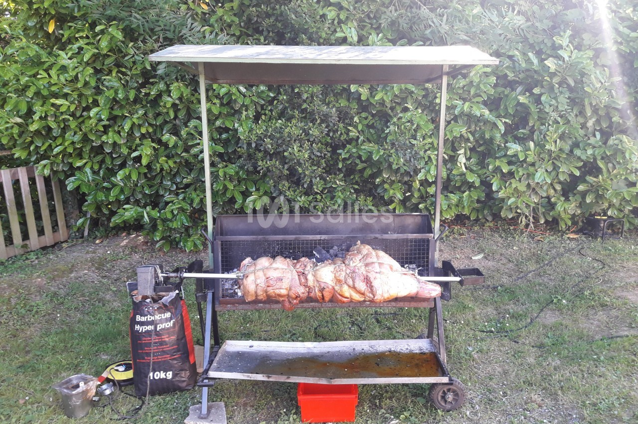 Un barbecue à rôtissoire en plein air avec des morceaux de viande en cuisson devant une haie verte.