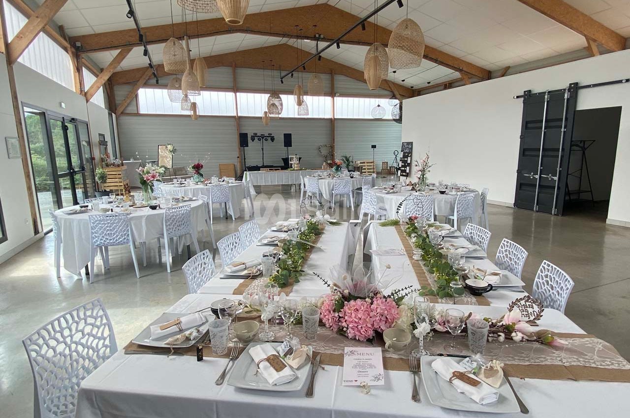 Salle de réception décorée pour un événement, avec tables dressées, fleurs et éclairage naturel.