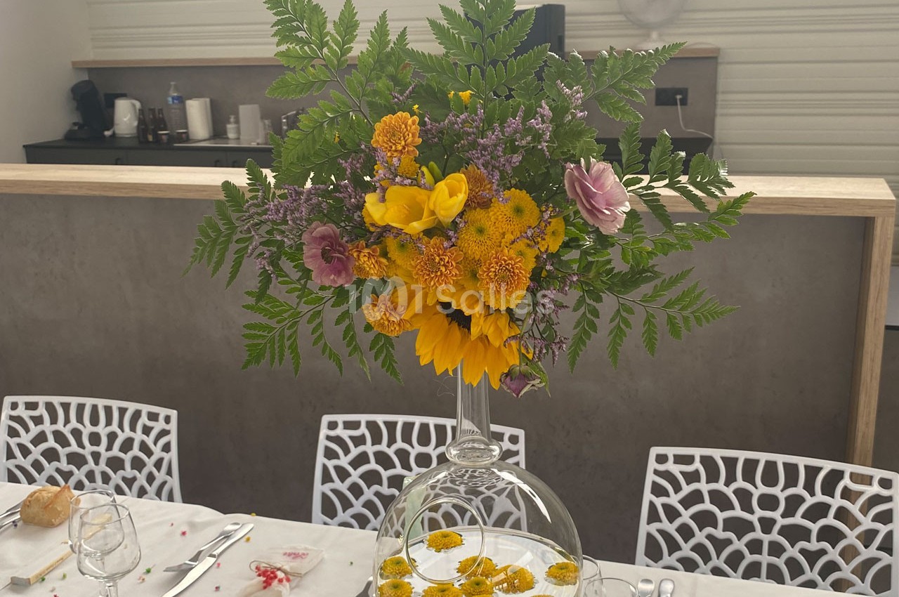 Centre de table floral avec tournesols, fougères et fleurs colorées, posé sur une table dressée dans une salle lumineuse.