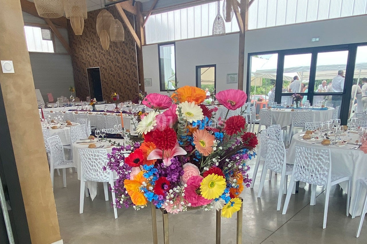 Composition florale colorée au premier plan dans une salle de réception lumineuse avec tables dressées en arrière-plan.