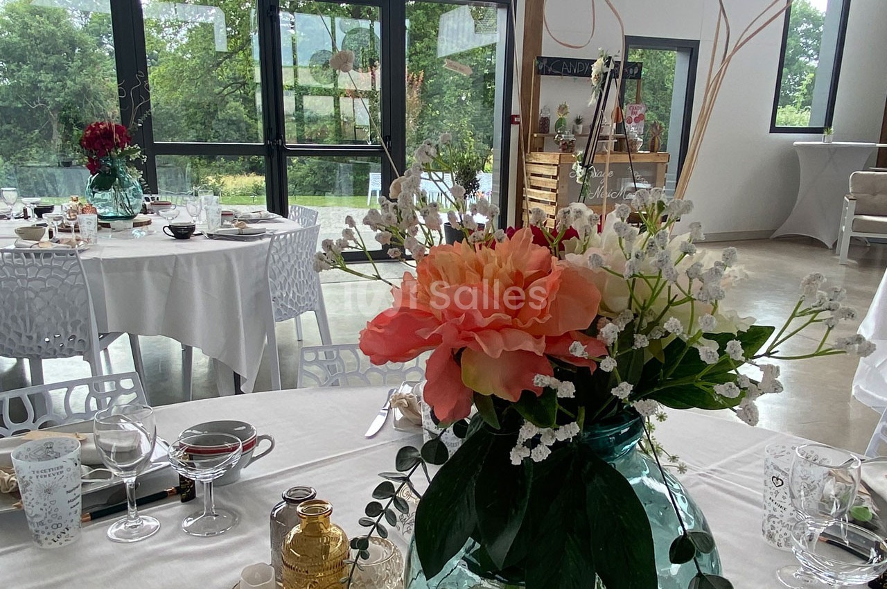 Salle de réception lumineuse avec tables décorées, centre de table floral et grandes baies vitrées donnant sur un jardin.