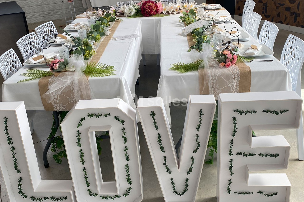 Table en U décorée pour un événement, ornée de fleurs, feuillages et lettres blanches formant le mot ’LOVE’.