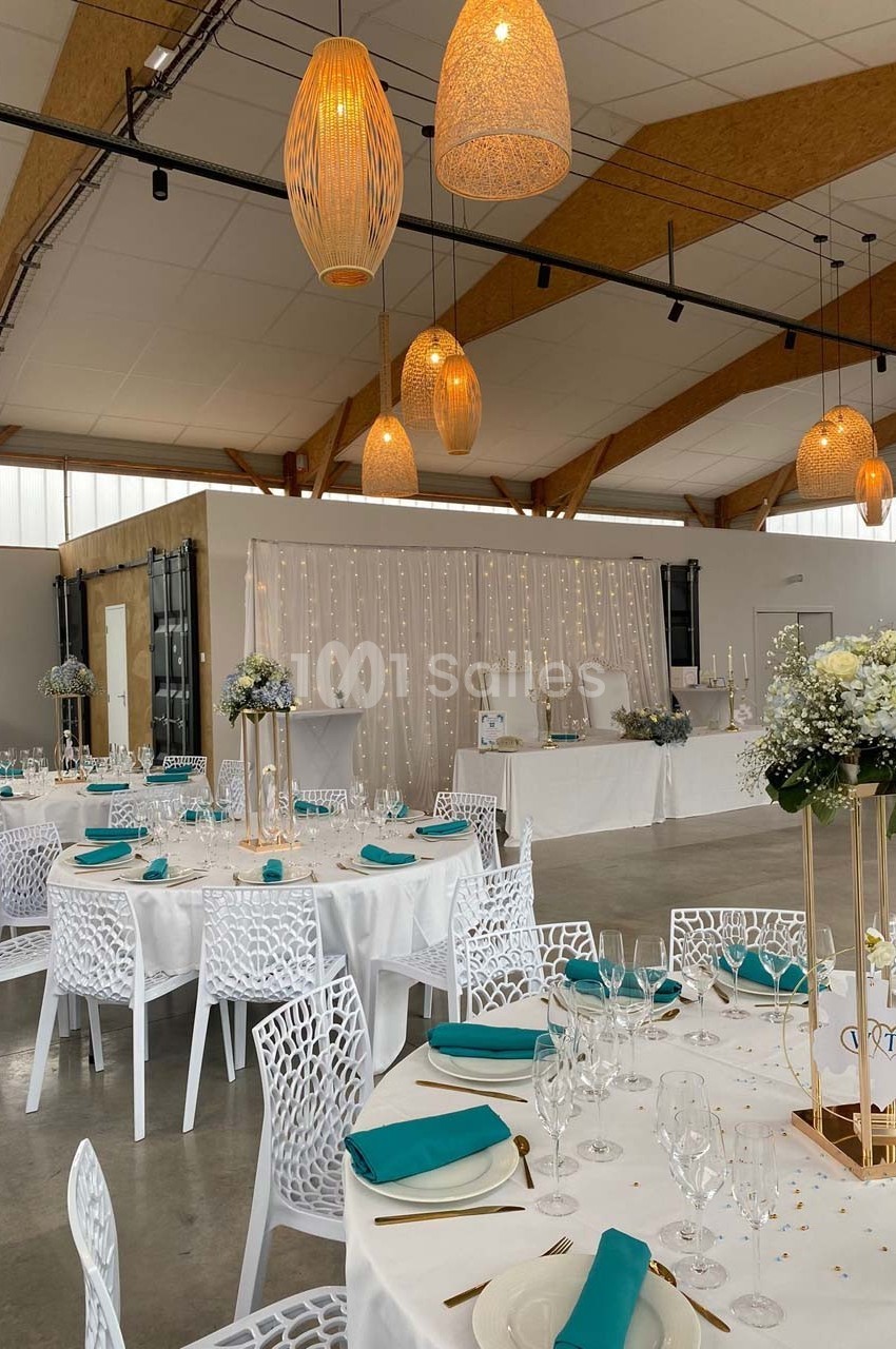 Salle de réception décorée avec des tables rondes, nappes blanches, serviettes turquoise et éclairage suspendu chaleureux.