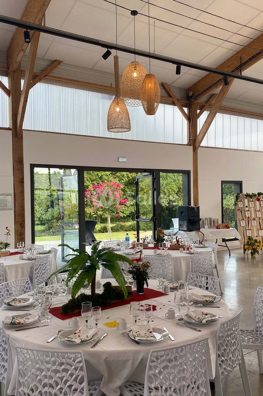 Salle lumineuse avec tables rondes dressées, décorations végétales et grandes baies vitrées donnant sur un jardin.