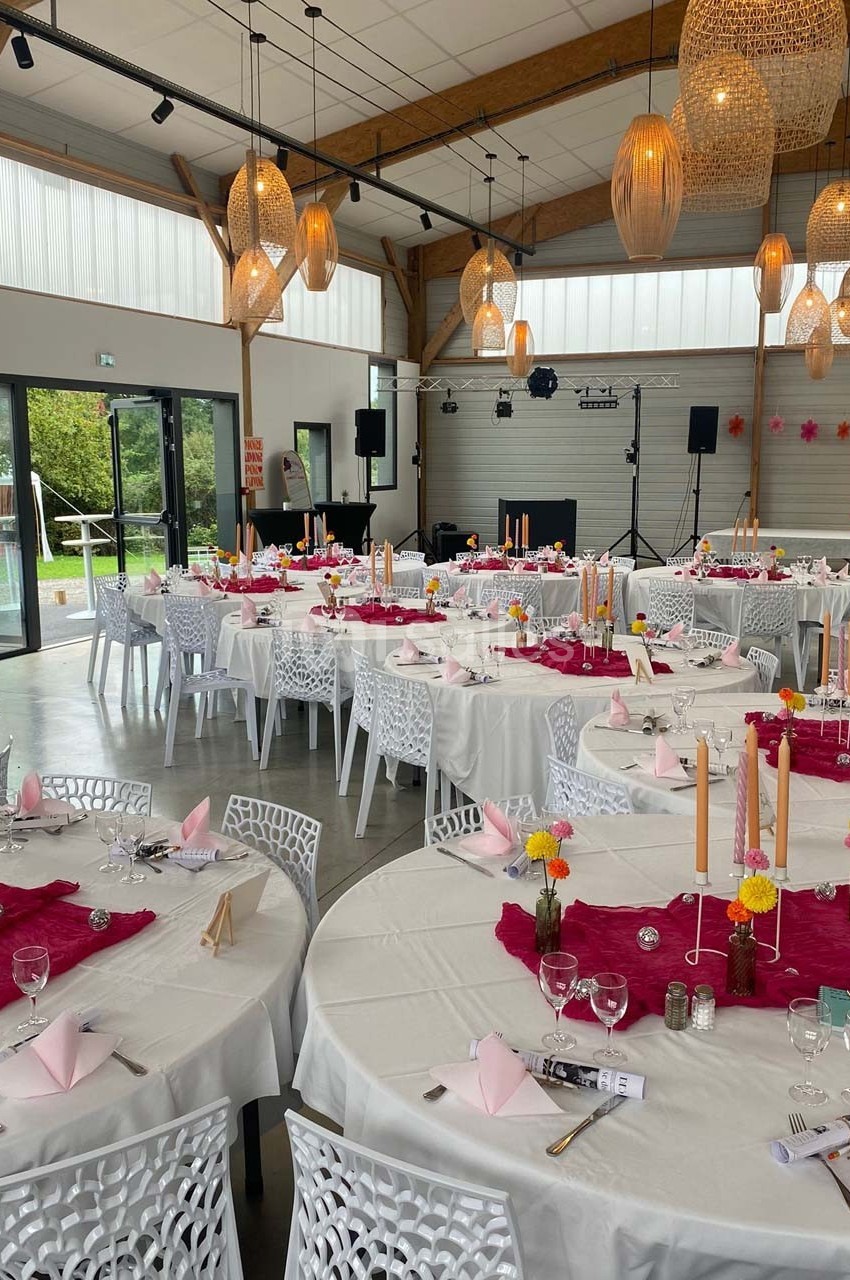 Salle de réception décorée avec des tables rondes, nappes blanches, chemins de table roses et chandeliers.