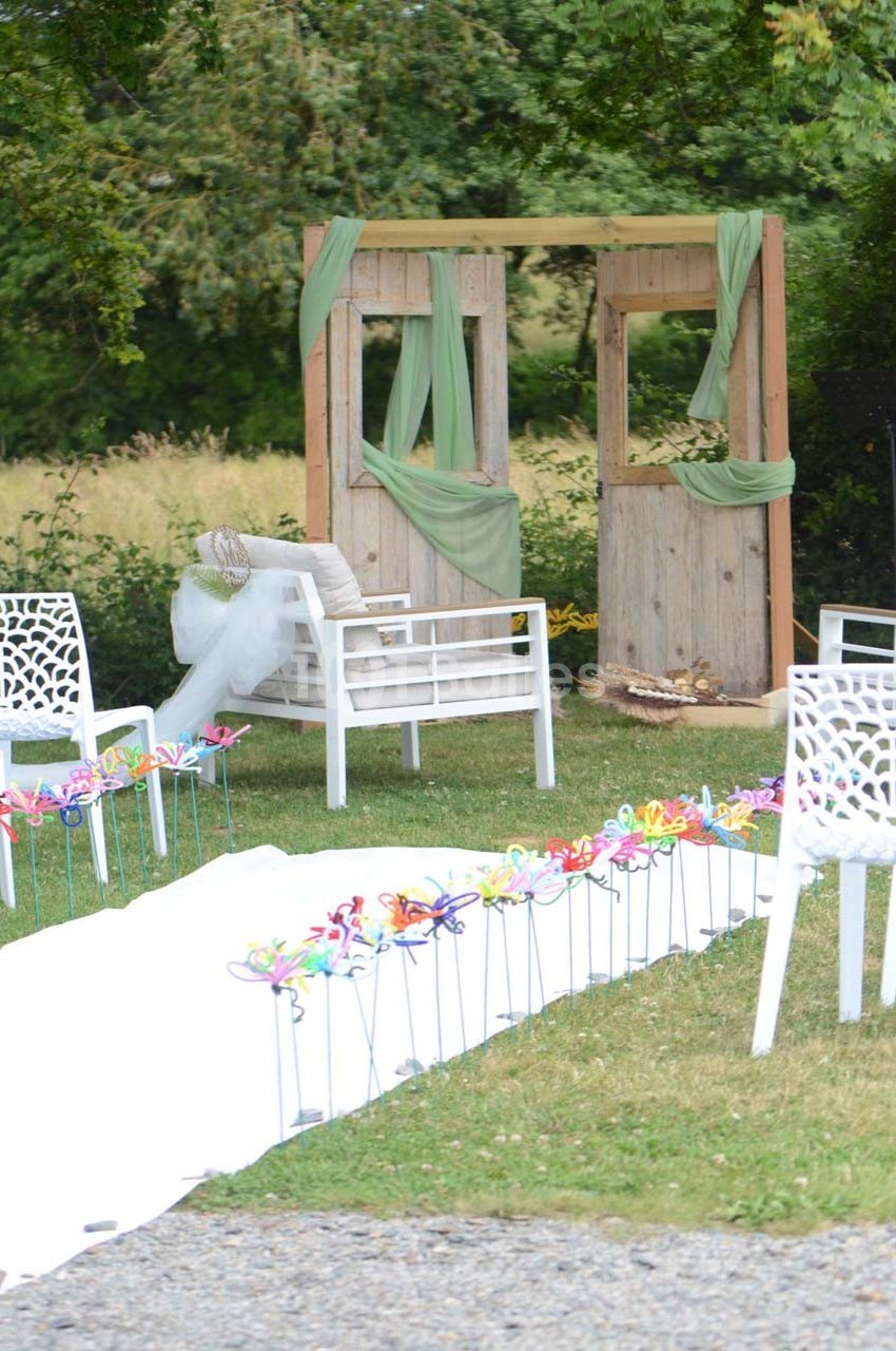 Décor champêtre avec arche en bois, chaises blanches et allée ornée de moulins à vent colorés sur pelouse.
