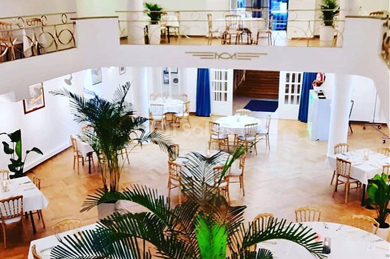 Salle lumineuse avec parquet, tables rondes dressées, chaises dorées et plantes vertes en décoration.