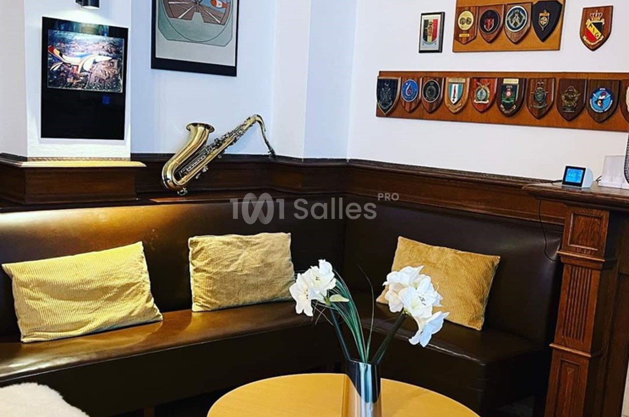 Coin salon avec banquette en cuir, coussins dorés, saxophone mural, décorations militaires et vase de fleurs blanches.