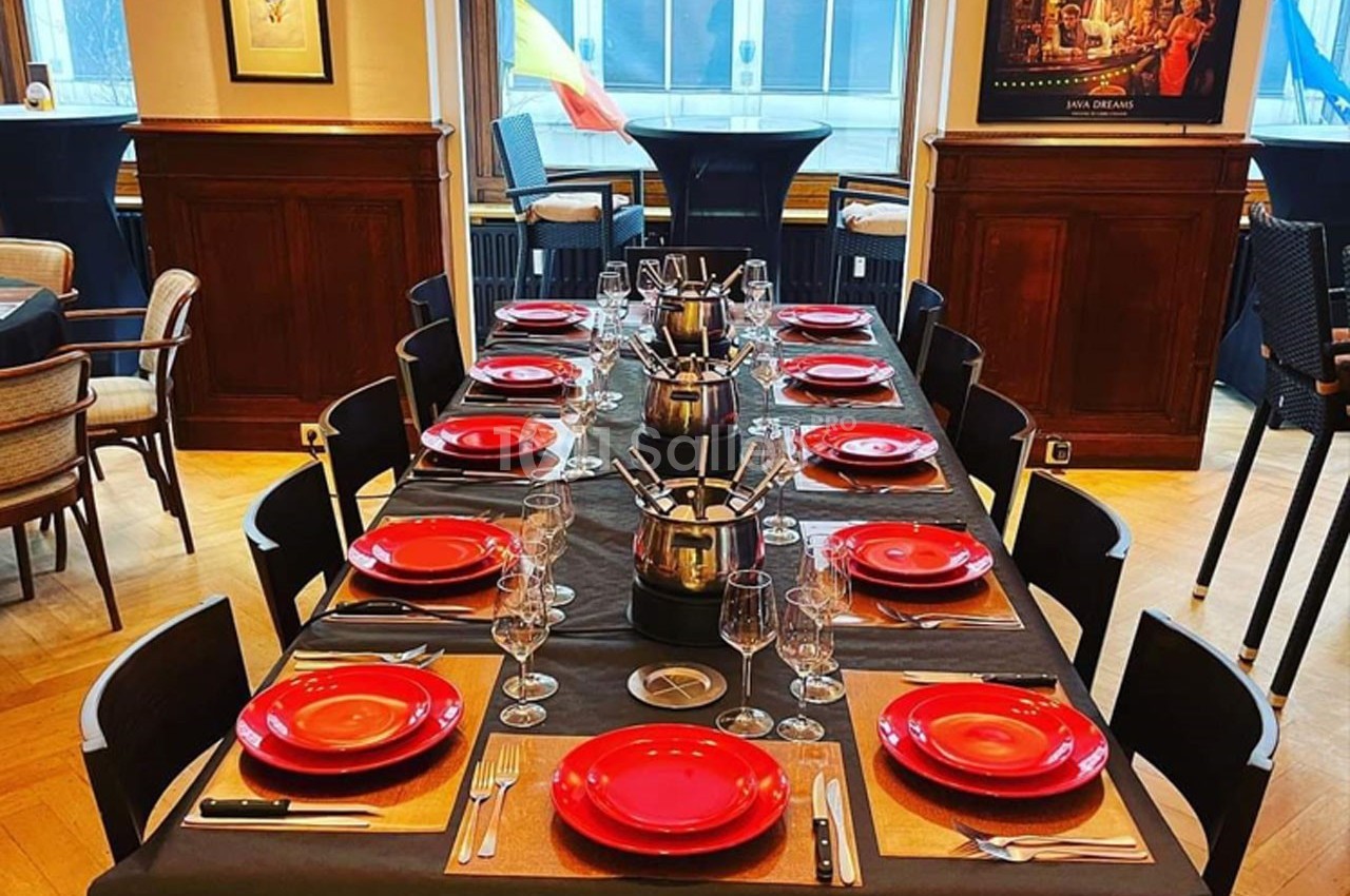 Table dressée avec des assiettes rouges, couverts et verres, entourée de chaises dans une salle lumineuse au décor…