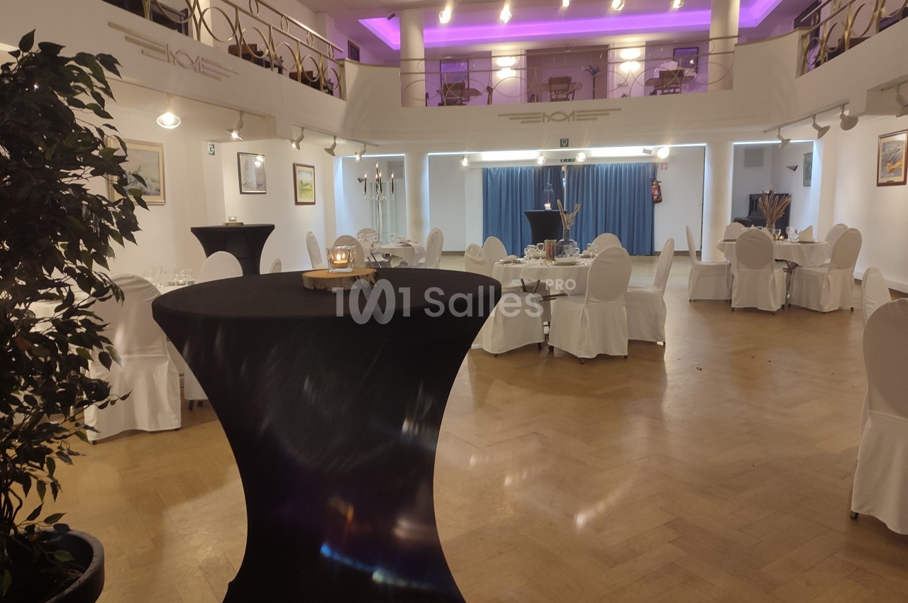 Salle de réception lumineuse avec tables rondes dressées, chaises blanches et éclairage violet au plafond.