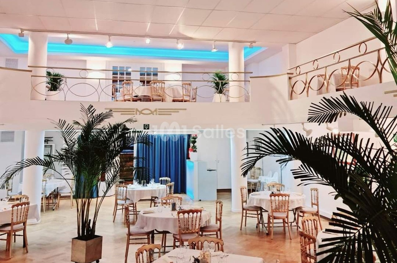 Salle lumineuse avec tables dressées, chaises en bois, plantes décoratives et mezzanine surplombant l'espace.