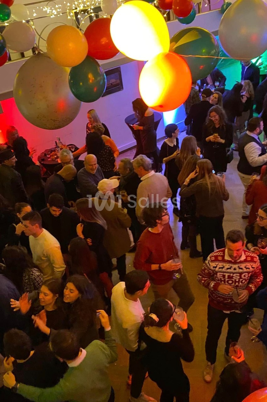 Groupe de personnes dans une salle festive décorée de ballons colorés, discutant et dansant lors d'un événement.