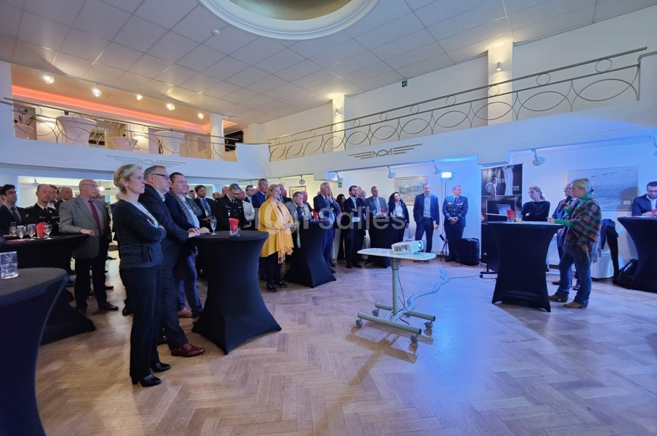 Groupe de personnes debout dans une salle lumineuse, participant à un événement avec des tables hautes et un projecteur.