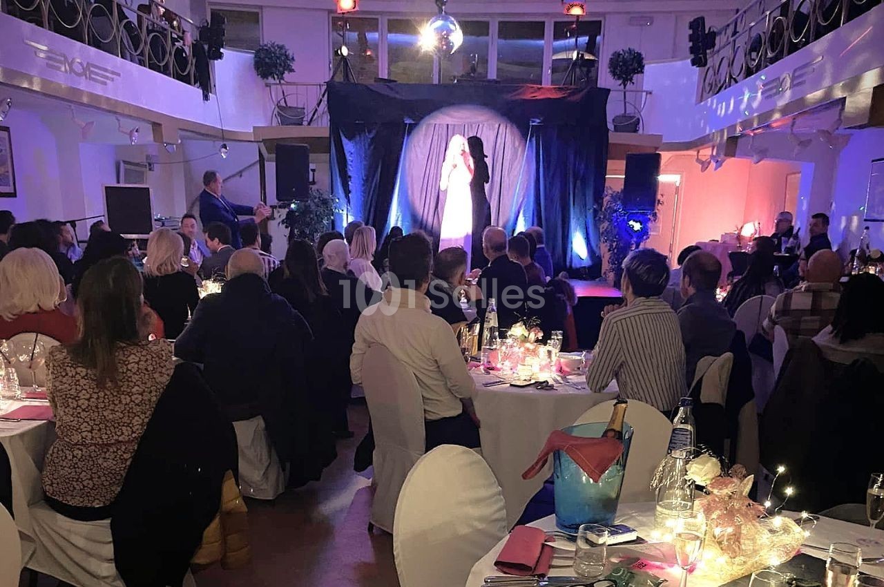 Une chanteuse sur scène éclairée, devant un public assis à des tables dans une salle de spectacle décorée.