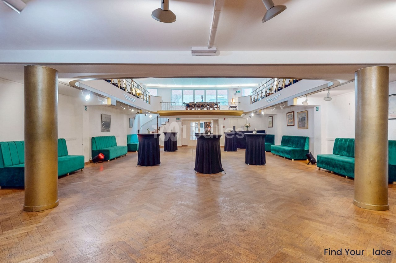 Salle spacieuse avec parquet en bois, colonnes dorées, canapés verts et tables hautes, éclairée par des fenêtres.