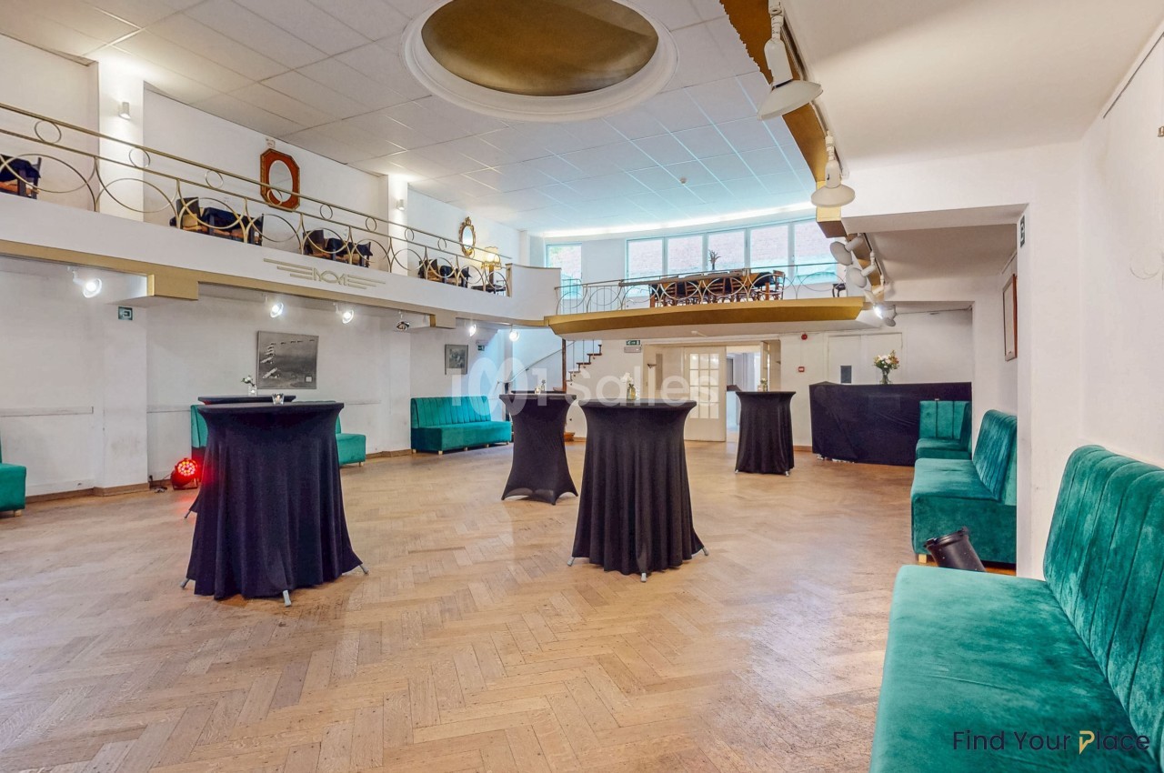 Salle lumineuse avec parquet en bois, tables hautes noires, banquettes vertes et mezzanine avec garde-corps doré.