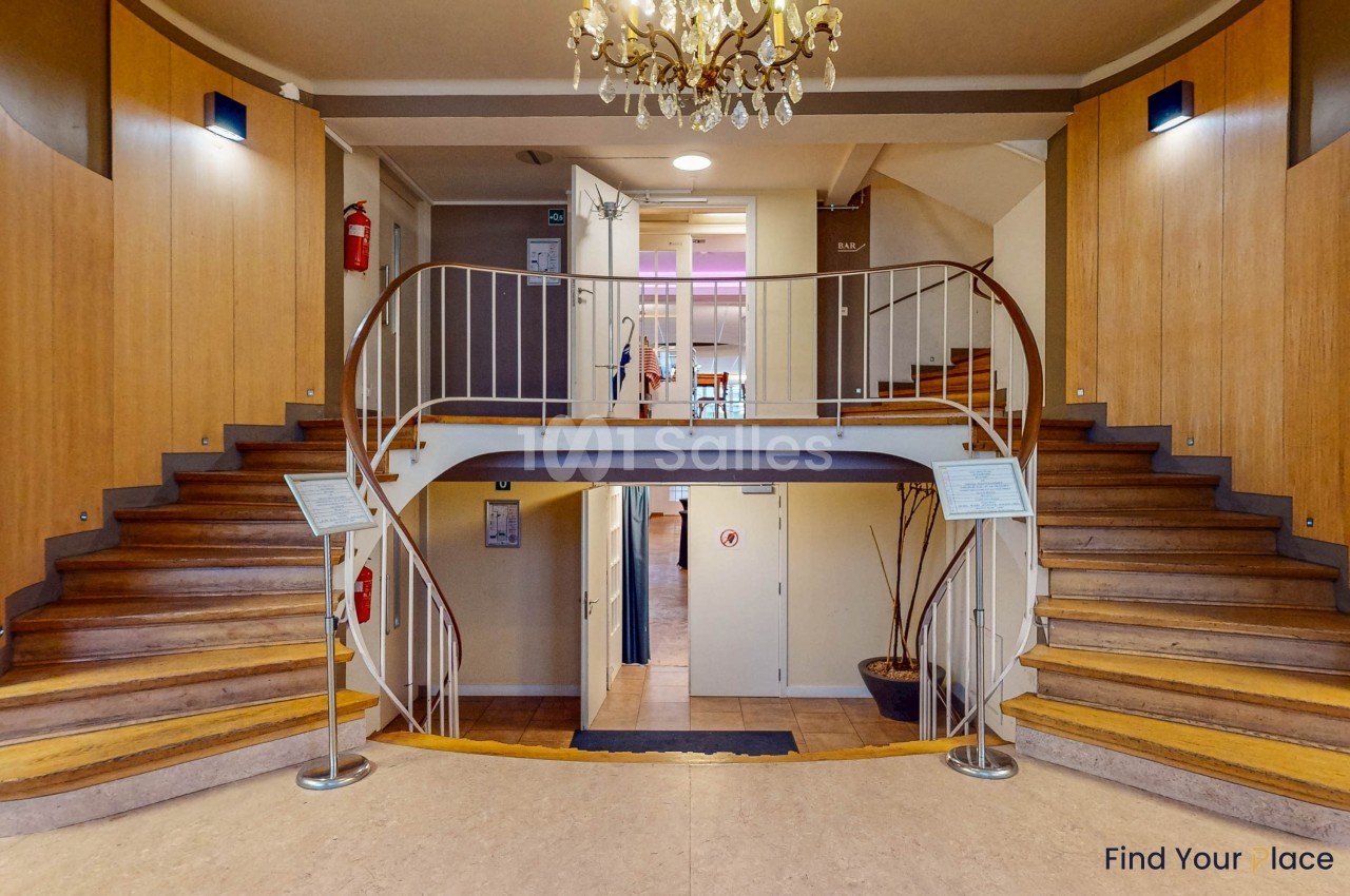 Escalier en bois à double volée dans un hall avec murs clairs, rampe métallique et lustre suspendu.