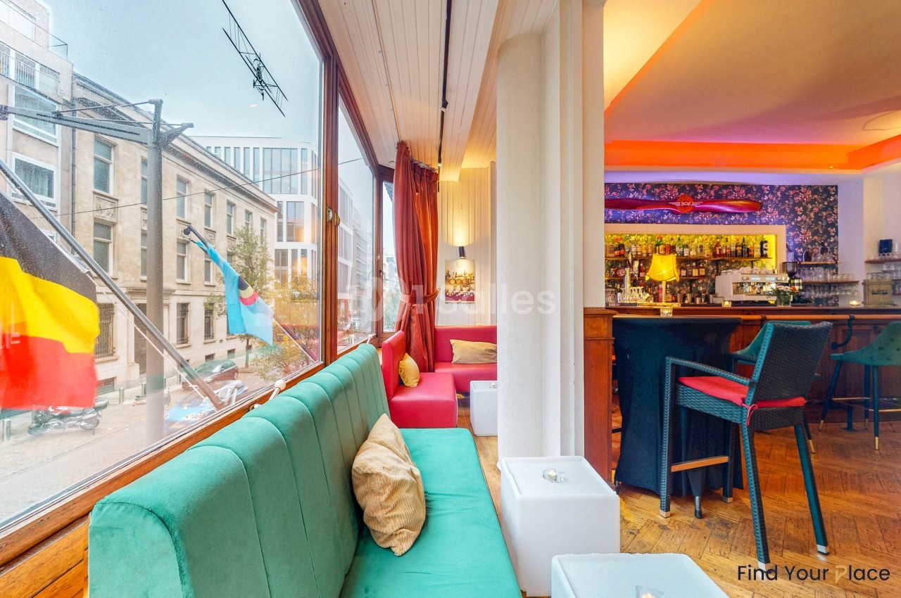 Intérieur d'un café lumineux avec canapés colorés, grandes fenêtres donnant sur une rue et un bar en arrière-plan.