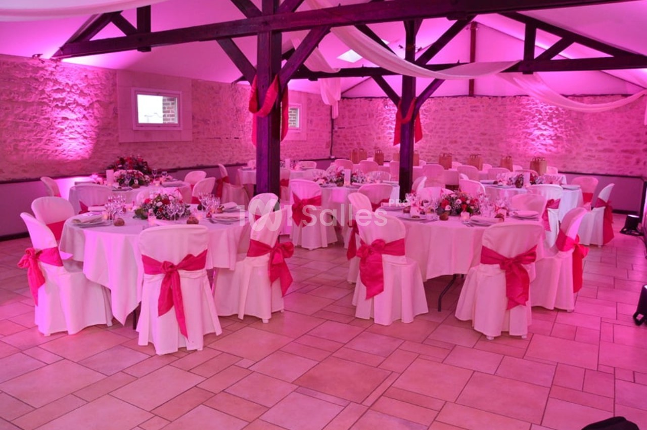Salle de réception décorée avec des tables rondes, nappes blanches, chaises ornées de nœuds roses et éclairage rose.