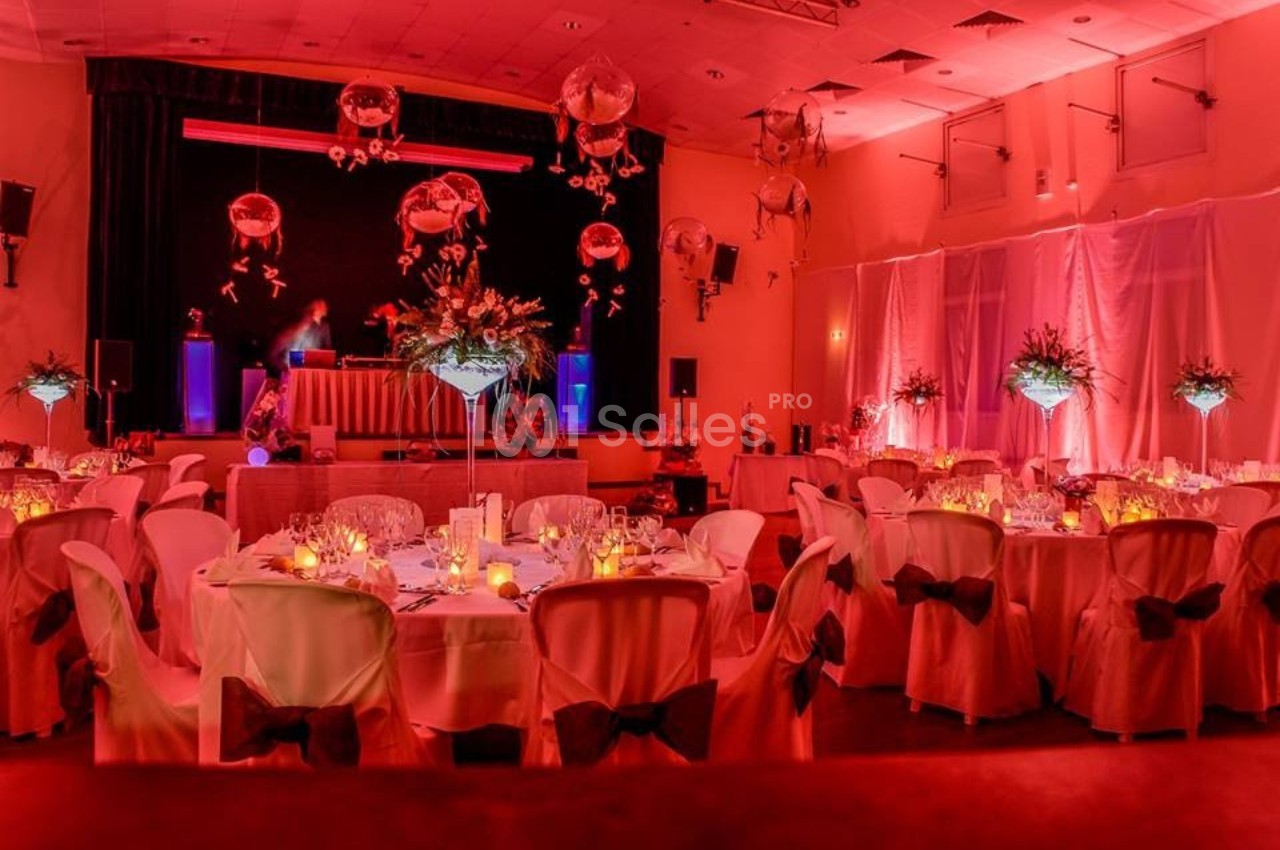 Salle de réception décorée avec des tables rondes, nappes blanches, éclairage rouge et arrangements floraux suspendus.