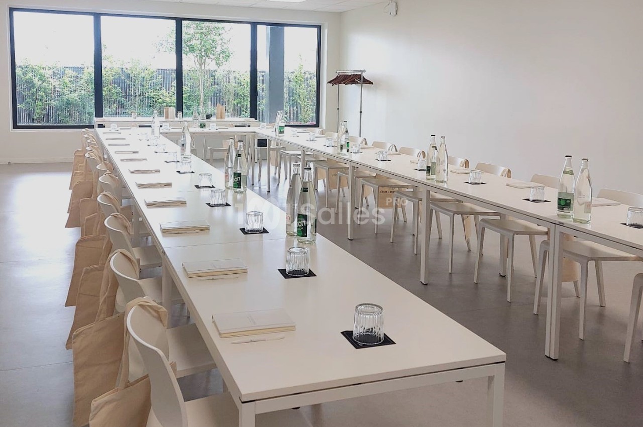 Salle lumineuse aménagée avec de longues tables blanches, des chaises, des bouteilles d'eau et des carnets disposés.