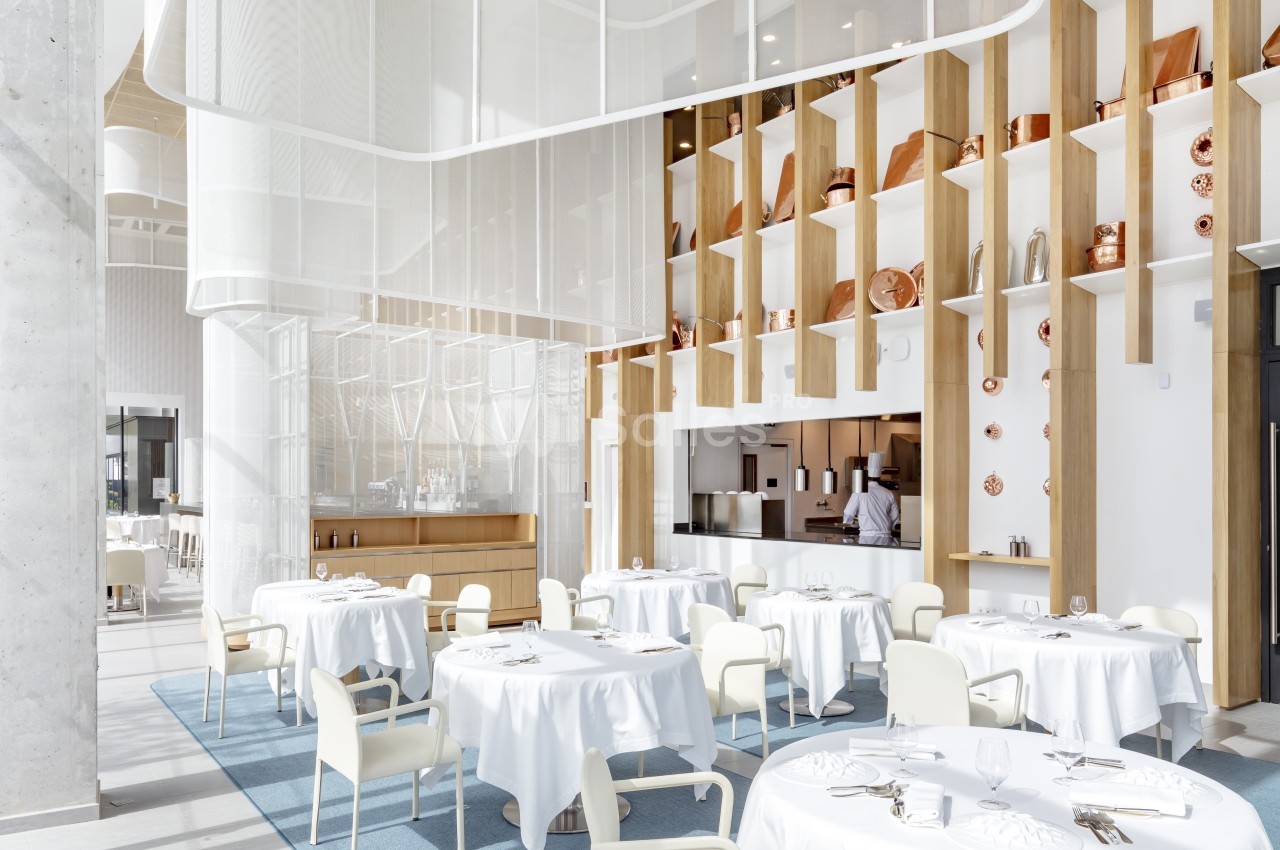 Salle de restaurant lumineuse avec tables dressées, chaises blanches et étagères décorées d'objets en cuivre.