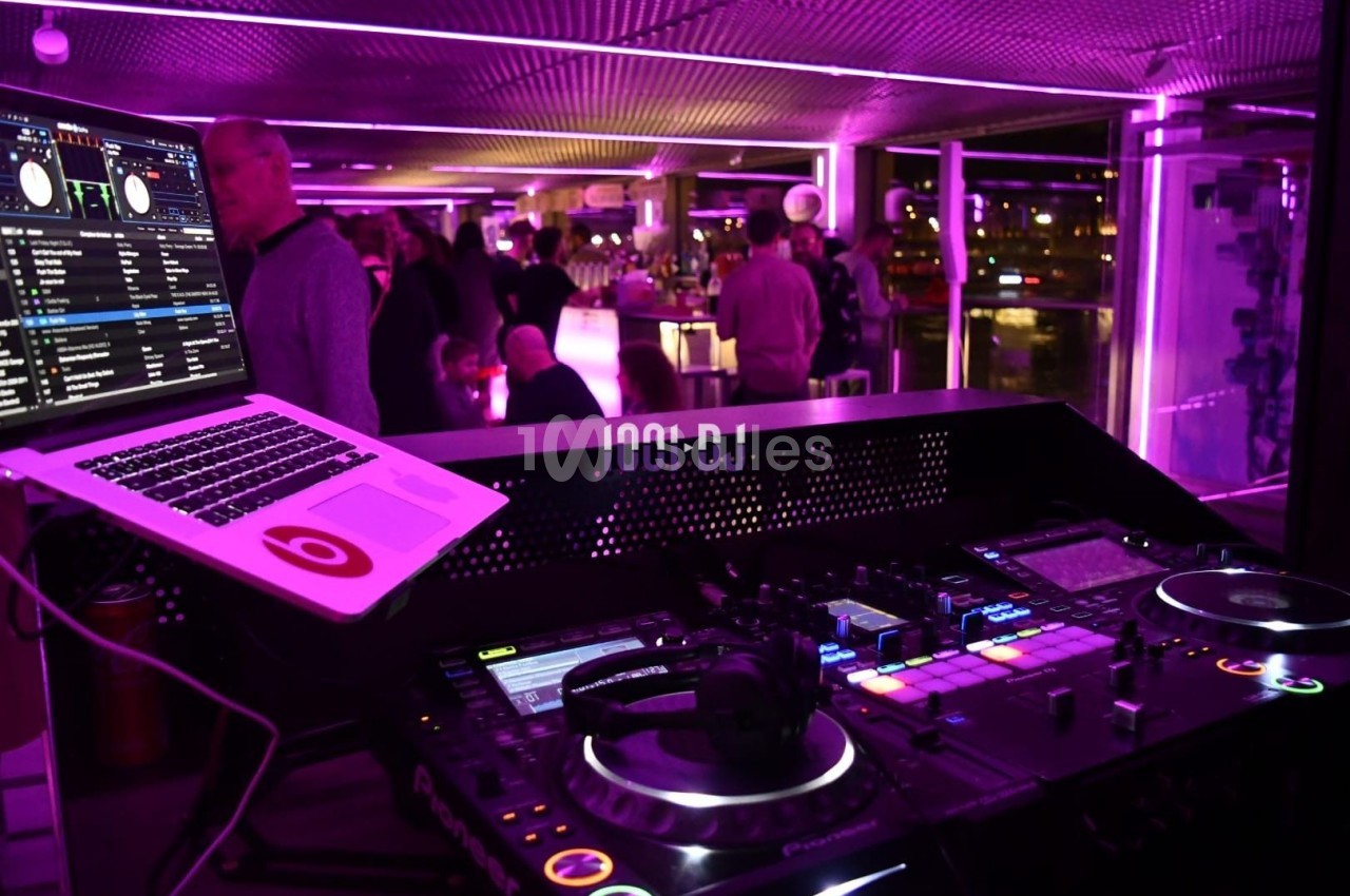 Poste de DJ avec platines et ordinateur dans un espace éclairé par des lumières violettes, avec des personnes en arrière…