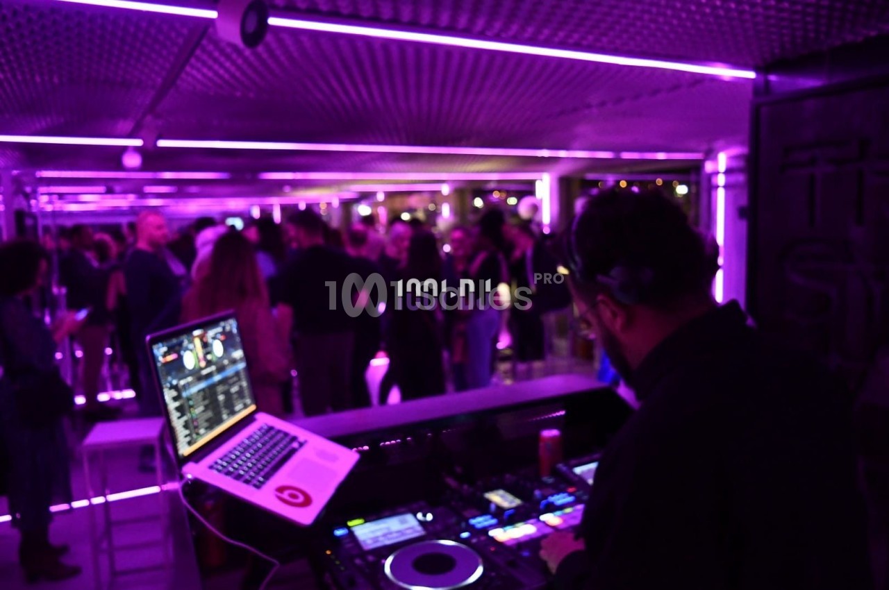 Un DJ mixe de la musique dans un club éclairé par des lumières violettes, avec une foule en arrière-plan.