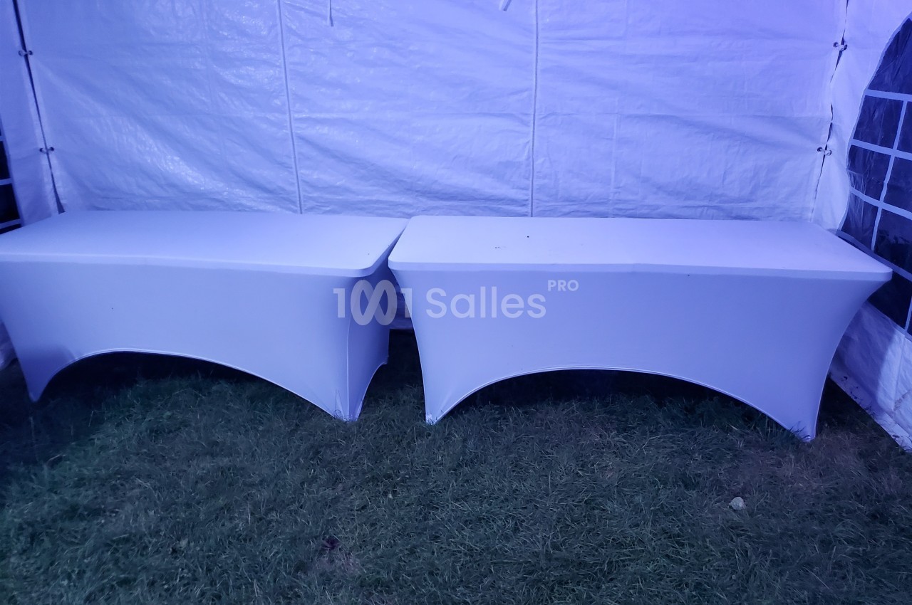 Deux tables rectangulaires recouvertes de housses blanches, placées côte à côte sur une pelouse sous une tente.