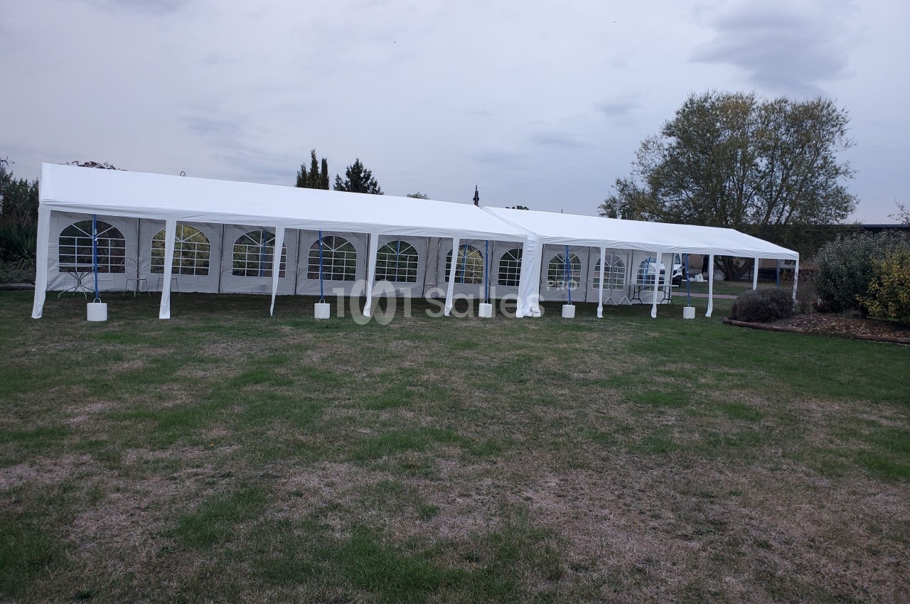 Grande tente blanche avec fenêtres transparentes installée sur une pelouse, entourée d'arbres et d'arbustes.